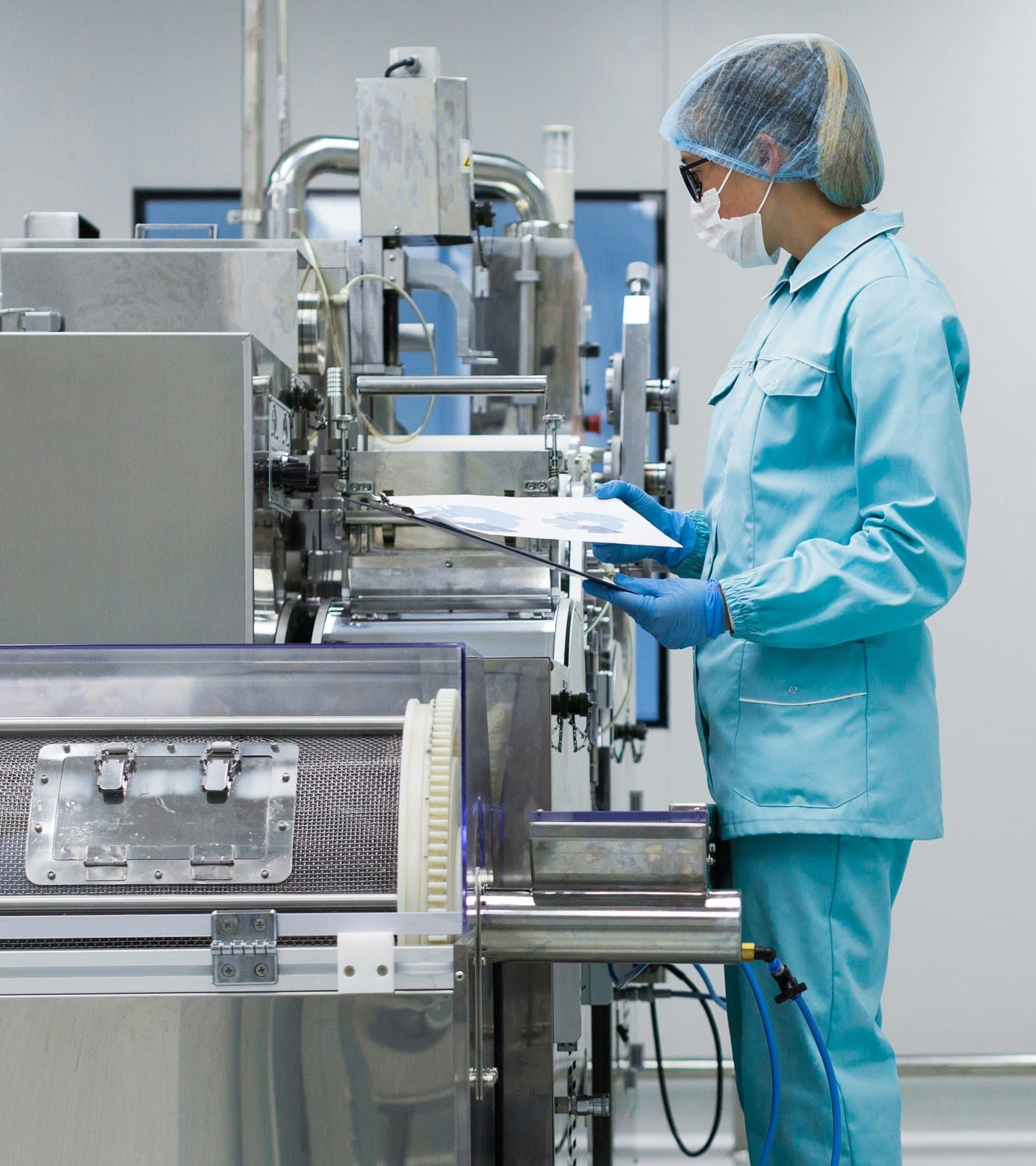 Technician in a cleanroom gown, hairnet, mask, and gloves checking a sheet on a clipboard at a stainless production machine
